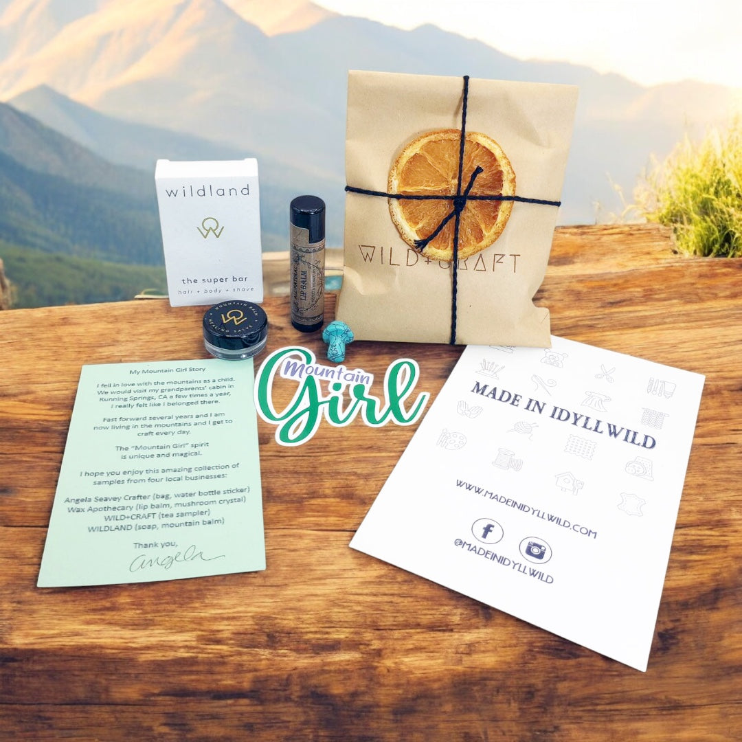 Mountain Girl Bundle contents, travel size super bar and mountain balm by Wildland, Small Turquoise stone mushroom,  Lip Balm by Wax Apothecary, Tea sampler envelope of 3  teas by Wild + Craft, Mountain Girl story card and Mountain Girl Sticker by Angela Seavey Crafter, and Made in Idyllwild post card, all laying on wood with mountain skyline in the background
