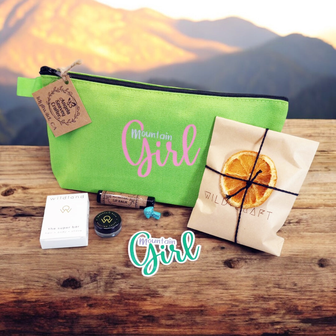 Mountain Girl Bundle green zipper bag with contents, travel size super bar and mountain balm by Wildland, Small Turquoise stone mushroom, Lip Balm by Wax Apothecary, Tea sampler envelope of 3 teas by Wild + Craft, Mountain Girl story card and Mountain Girl Sticker by Angela Seavey Crafter, and Made in Idyllwild post card, all laying on wood with mountain skyline in the background