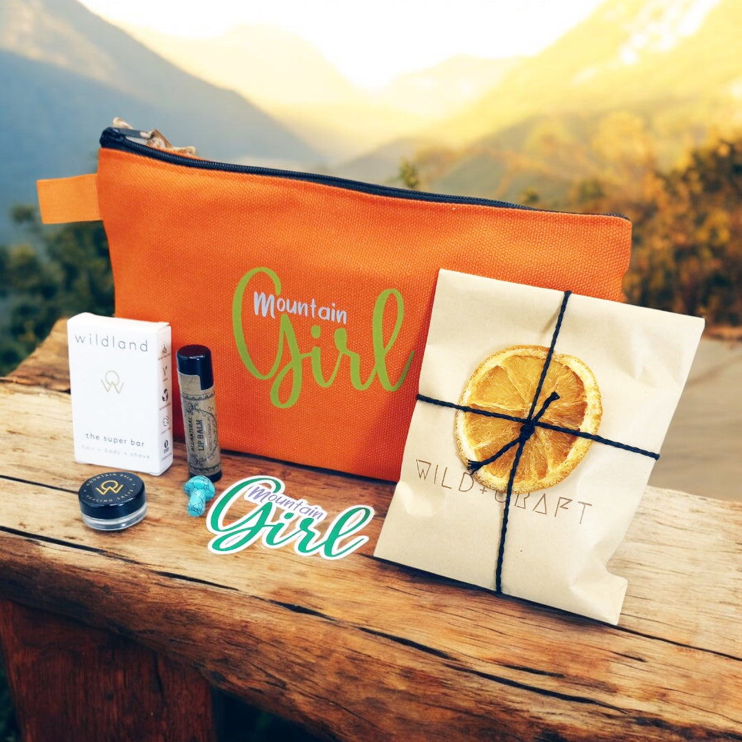Mountain Girl Bundle orange zipper bag with contents, travel size super bar and mountain balm by Wildland, Small Turquoise stone mushroom, Lip Balm by Wax Apothecary, Tea sampler envelope of 3 teas by Wild + Craft, Mountain Girl story card and Mountain Girl Sticker by Angela Seavey Crafter, and Made in Idyllwild post card, all laying on wood with mountain skyline in the background