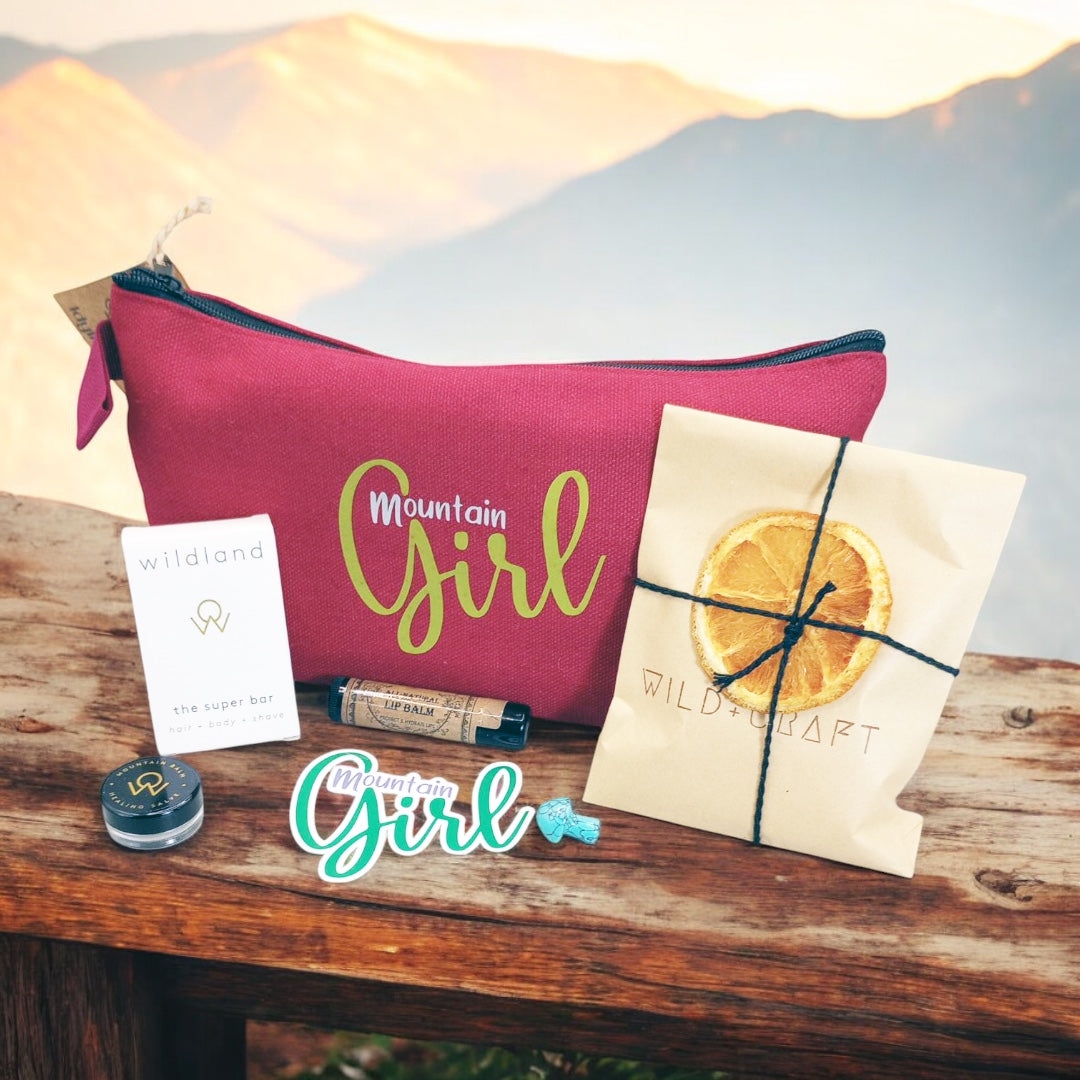 Mountain Girl Bundle red zipper bag with contents, travel size super bar and mountain balm by Wildland, Small Turquoise stone mushroom, Lip Balm by Wax Apothecary, Tea sampler envelope of 3 teas by Wild + Craft, Mountain Girl story card and Mountain Girl Sticker by Angela Seavey Crafter, and Made in Idyllwild post card, all laying on wood with mountain skyline in the background