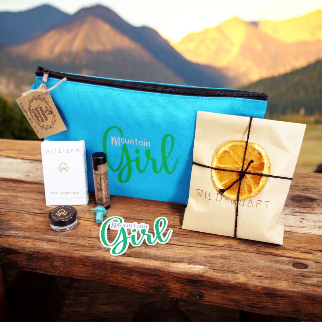 Mountain Girl Bundle turquoise blue zipper bag with contents, travel size super bar and mountain balm by Wildland, Small Turquoise stone mushroom, Lip Balm by Wax Apothecary, Tea sampler envelope of 3 teas by Wild + Craft, Mountain Girl story card and Mountain Girl Sticker by Angela Seavey Crafter, and Made in Idyllwild post card, all laying on wood with mountain skyline in the background