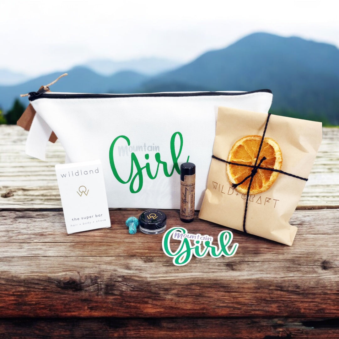 Mountain Girl Bundle white zipper bag with contents, travel size super bar and mountain balm by Wildland, Small Turquoise stone mushroom, Lip Balm by Wax Apothecary, Tea sampler envelope of 3 teas by Wild + Craft, Mountain Girl story card and Mountain Girl Sticker by Angela Seavey Crafter, and Made in Idyllwild post card, all laying on wood with mountain skyline in the background