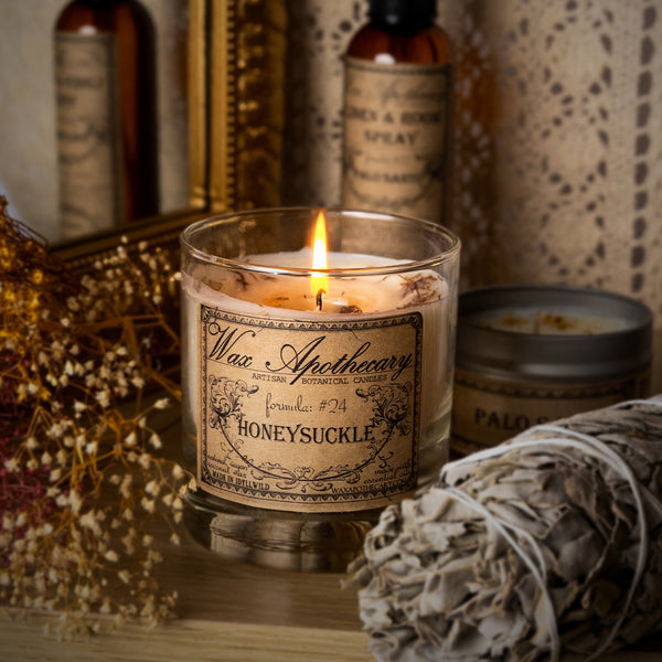 A lit candle in a clear glass jar with a vintage-style label reading 'Wax Apothecary' and 'HONEYSUCKLE', styled on a tabletop with dried flowers, a sage bundle, and other apothecary-style bottles in the background.