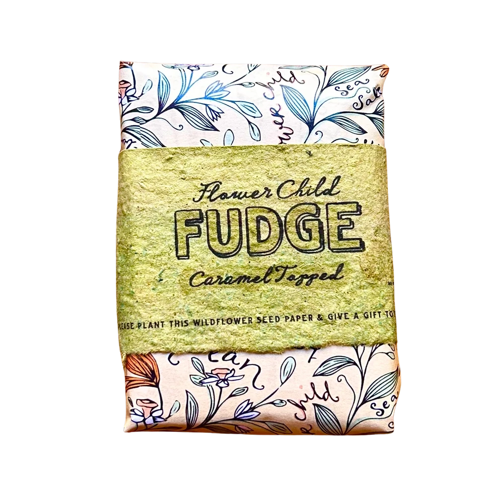 A square seed paper gift wrap featuring a central kraft paper band that reads 'Flower Child FUDGE Caramel Topped'. The top and bottom sections of the wrap are cream-colored with blue botanical illustrations and decorative text.
