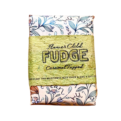 A square seed paper gift wrap featuring a central kraft paper band that reads 'Flower Child FUDGE Caramel Topped'. The top and bottom sections of the wrap are cream-colored with blue botanical illustrations and decorative text.