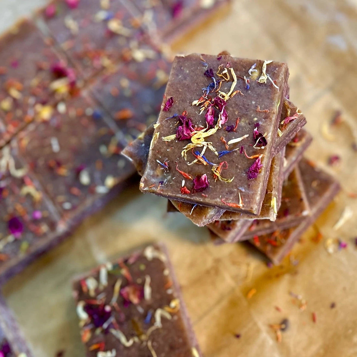 Dark chocolate fudge bars topped with dried flower petals, stacked on parchment paper with more bars blurred in the background.