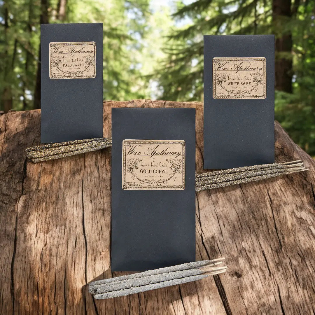 three envelopes of hand-rolled incense with incense sticks laying in front of them on a rustic wood slab with a forest landscape in the background 