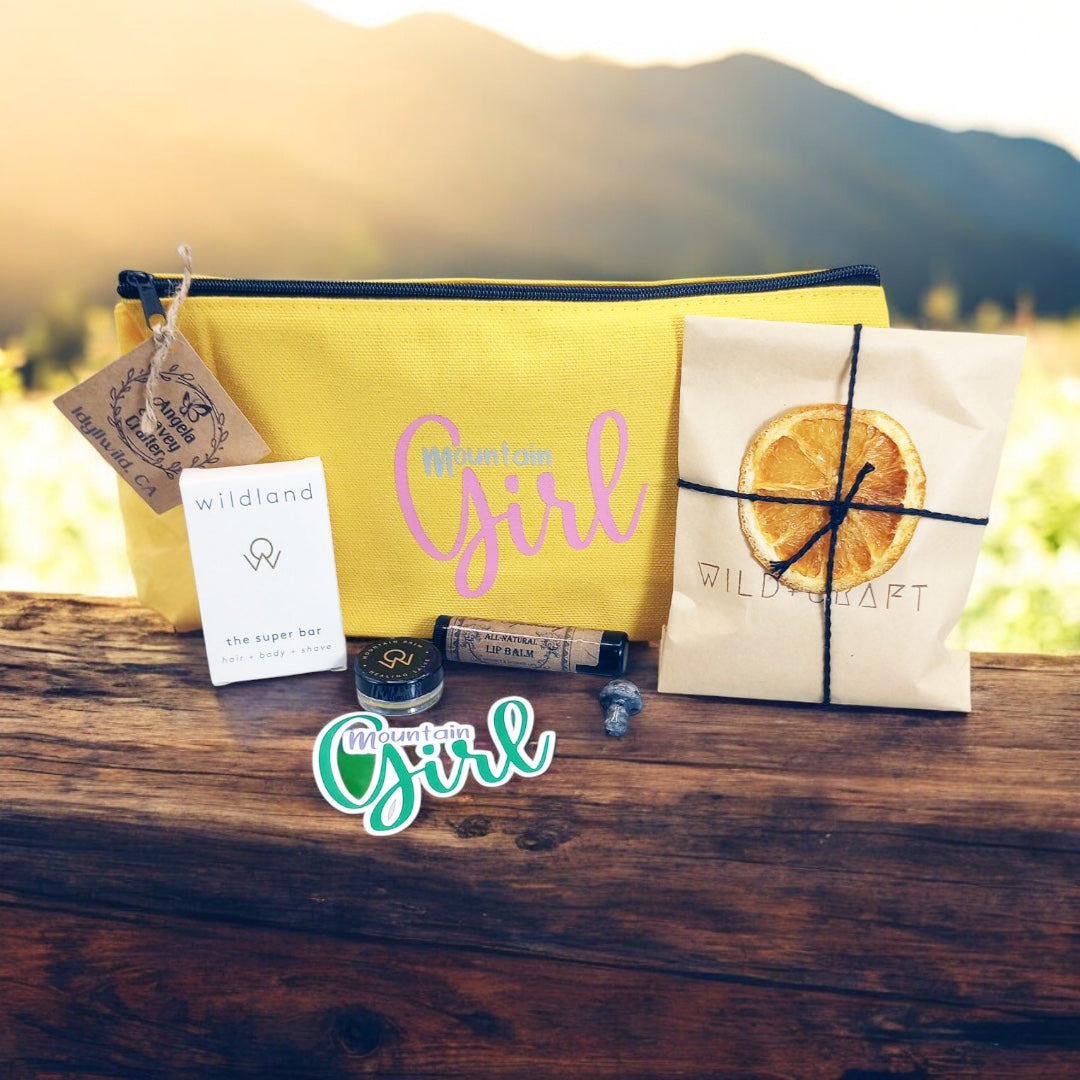 Mountain Girl Bundle yellow zipper bag with contents, travel size super bar and mountain balm by Wildland, Small Turquoise stone mushroom,  Lip Balm by Wax Apothecary, Tea sampler envelope of 3  teas by Wild + Craft, Mountain Girl story card and Mountain Girl Sticker by Angela Seavey Crafter, and Made in Idyllwild post card, all laying on wood with mountain skyline in the background