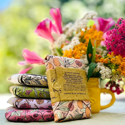 A stack of colorful reusable cloth napkins with botanical patterns, wrapped in a kraft paper band with printed text, displayed in front of a yellow mug filled with vibrant flowers against a soft green outdoor background.