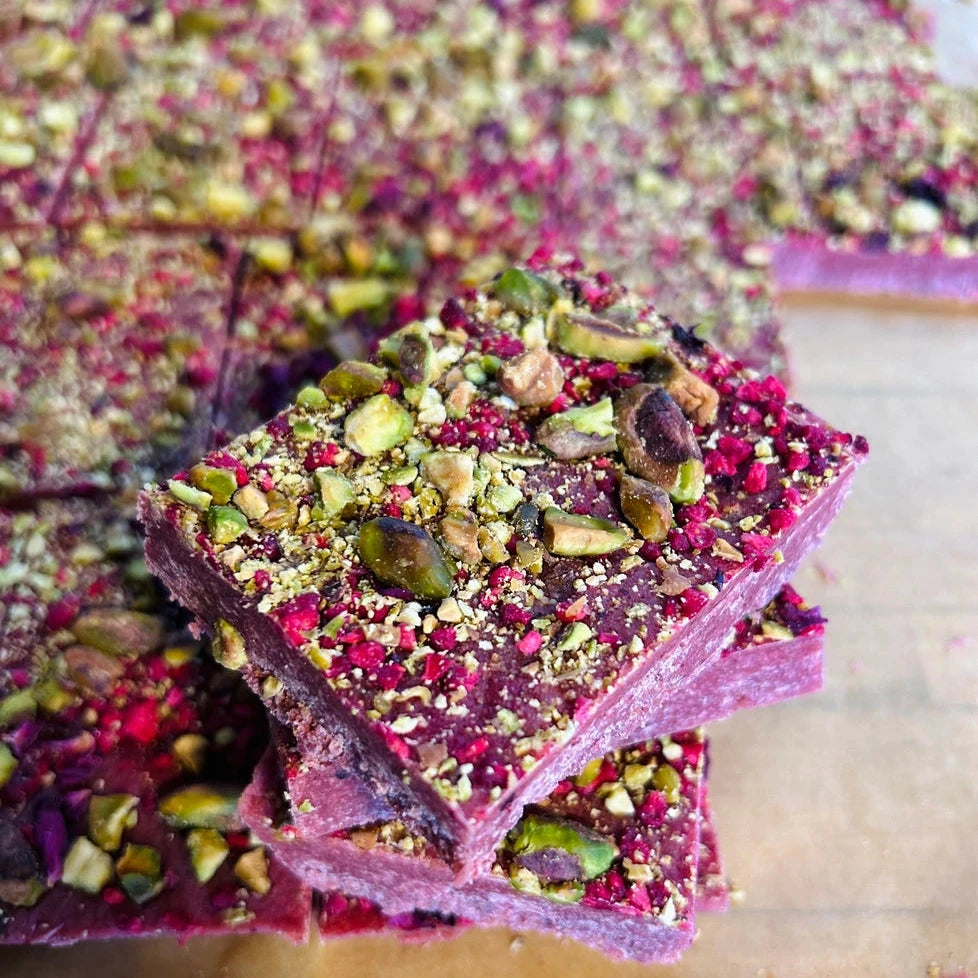 Close-up of square fudge dessert bars topped with chopped pistachios and pink dried flower pieces, arranged on parchment paper.