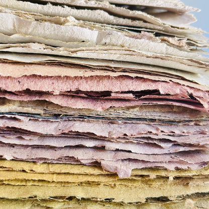 A close-up stack of textured, handmade paper sheets in various colors including cream, pink, purple, and yellow, stacked on a wooden surface.