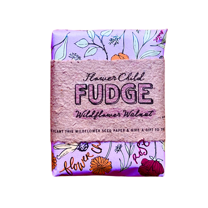 A packaged bar of Flower Child Fudge Wildflower Walnut fudge, wrapped in a textured pink paper band with floral illustrations on a light purple background.