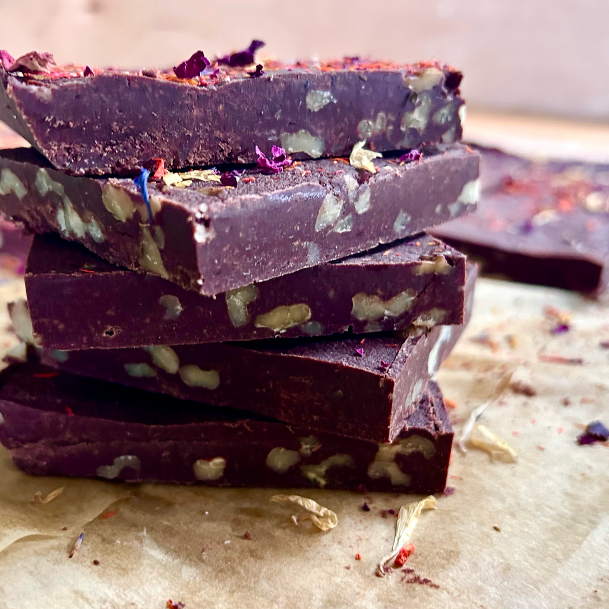 A stack of dark chocolate bark bars with chopped nuts and dried flower petals, photographed on parchment paper with more bars blurred in the background.